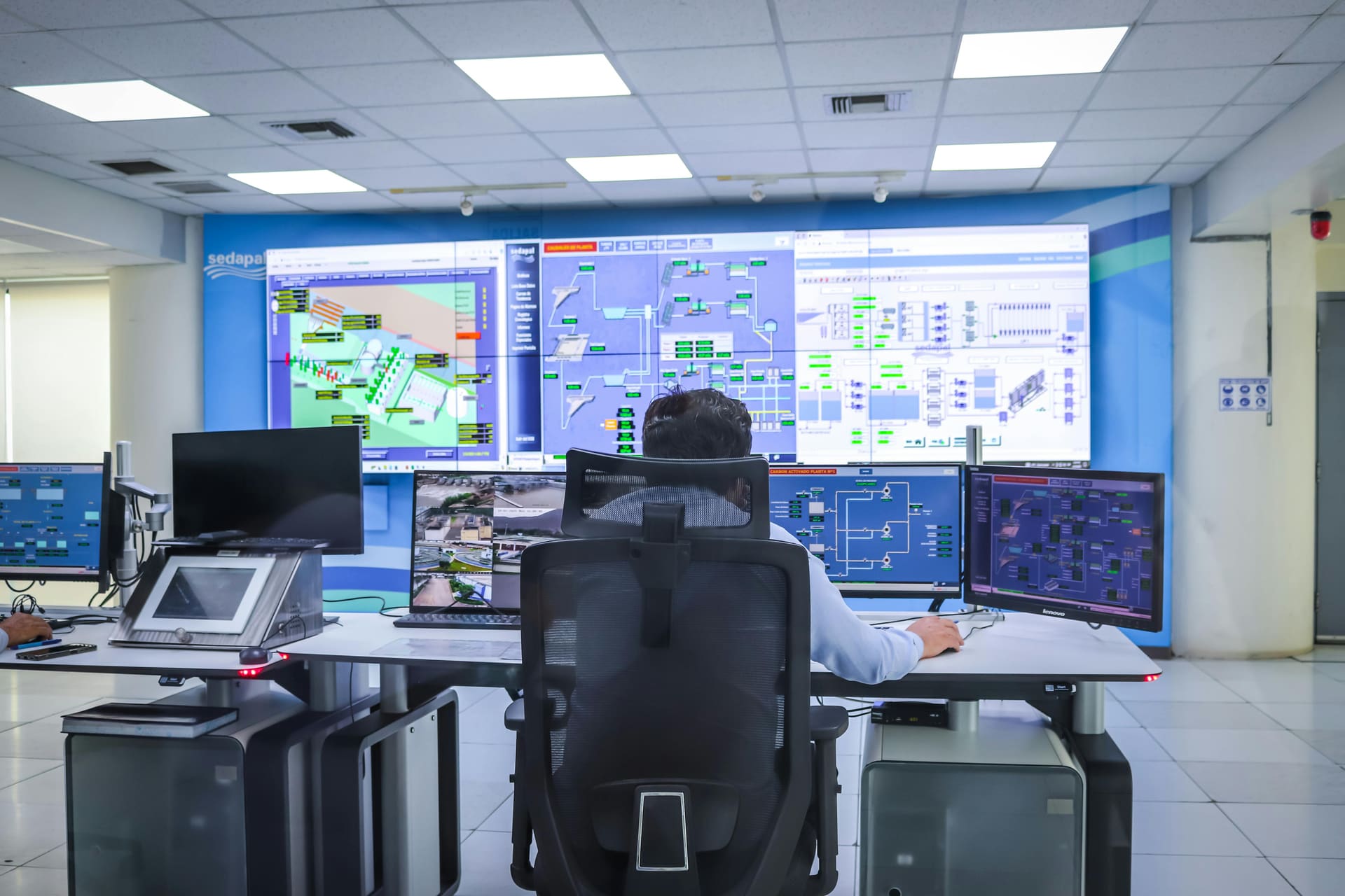 Operator seated at an industrial control room console with multiple SCADA monitoring screens displaying process schematics.