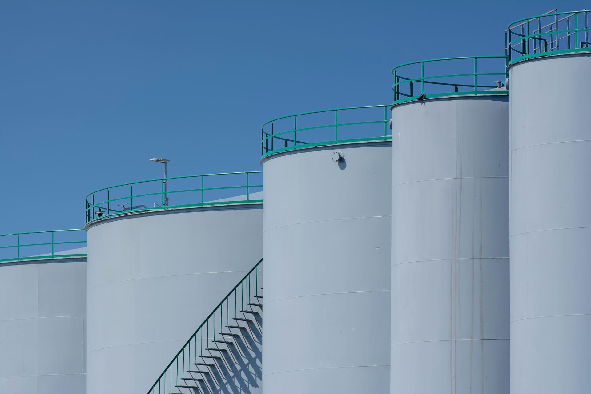 Row of clean white petroleum storage tanks with green safety railings under a clear blue sky.