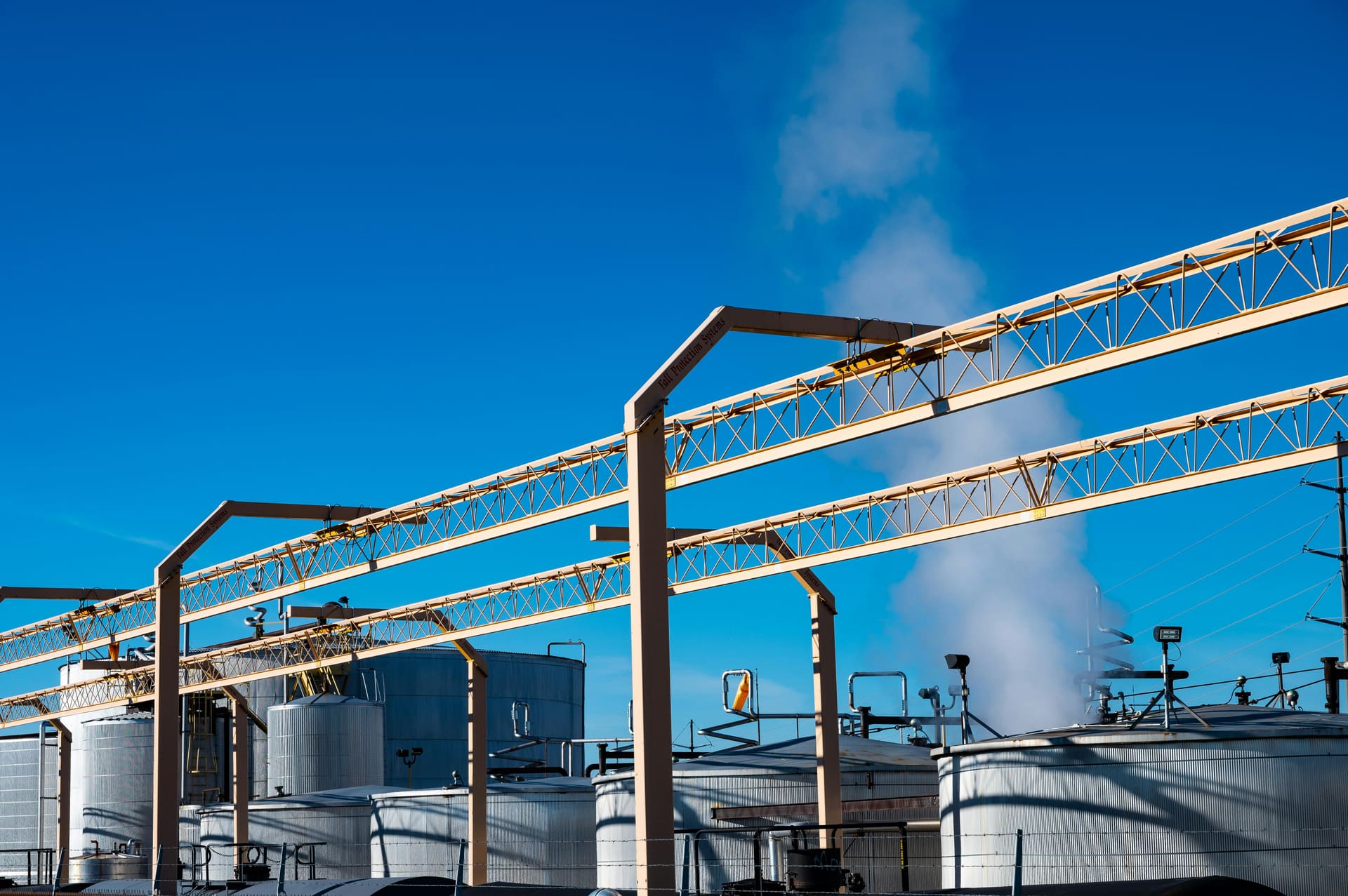 Petroleum storage tank farm with steel pipework and venting steam under a bright blue sky.