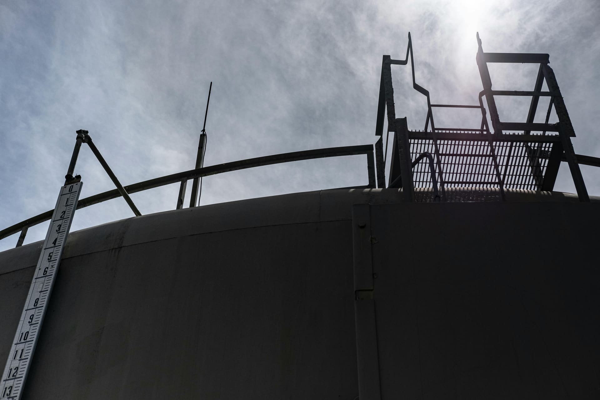 Silhouetted catwalk and stairs on the roof of a large petroleum storage tank against an overcast sky.