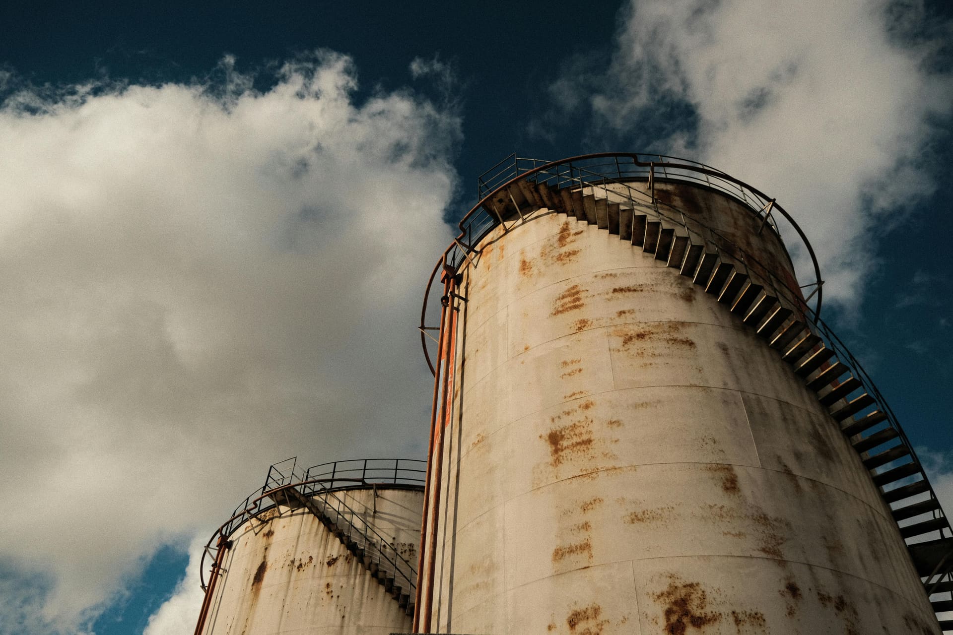 Two weathered petroleum storage tanks with spiral access stairs photographed from below against a dramatic cloudy sky.
